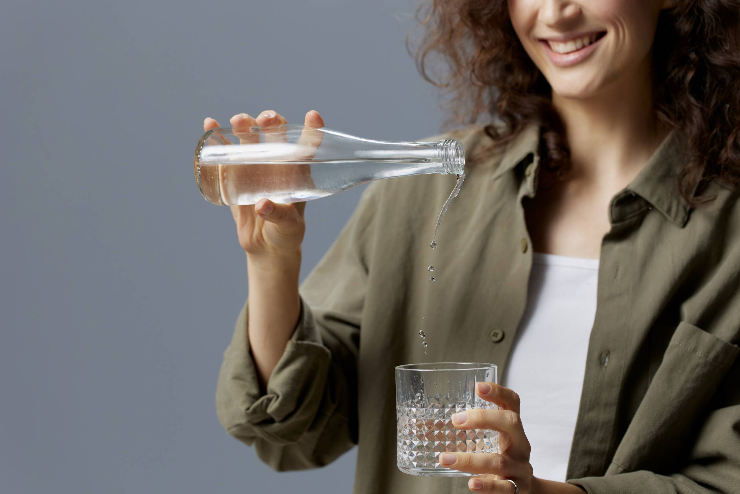 Happy smiling curly beautiful woman in casual khaki green shirt pours water from bottle in glass posing isolated on over gray blue background. Healthy lifestyle. Water is Life concept. Free place Happy smiling curly beautiful woman in casual khaki green shirt pours water from bottle in glass posing isolated on over gray blue background. Healthy lifestyle. Water is Life concept. Free place